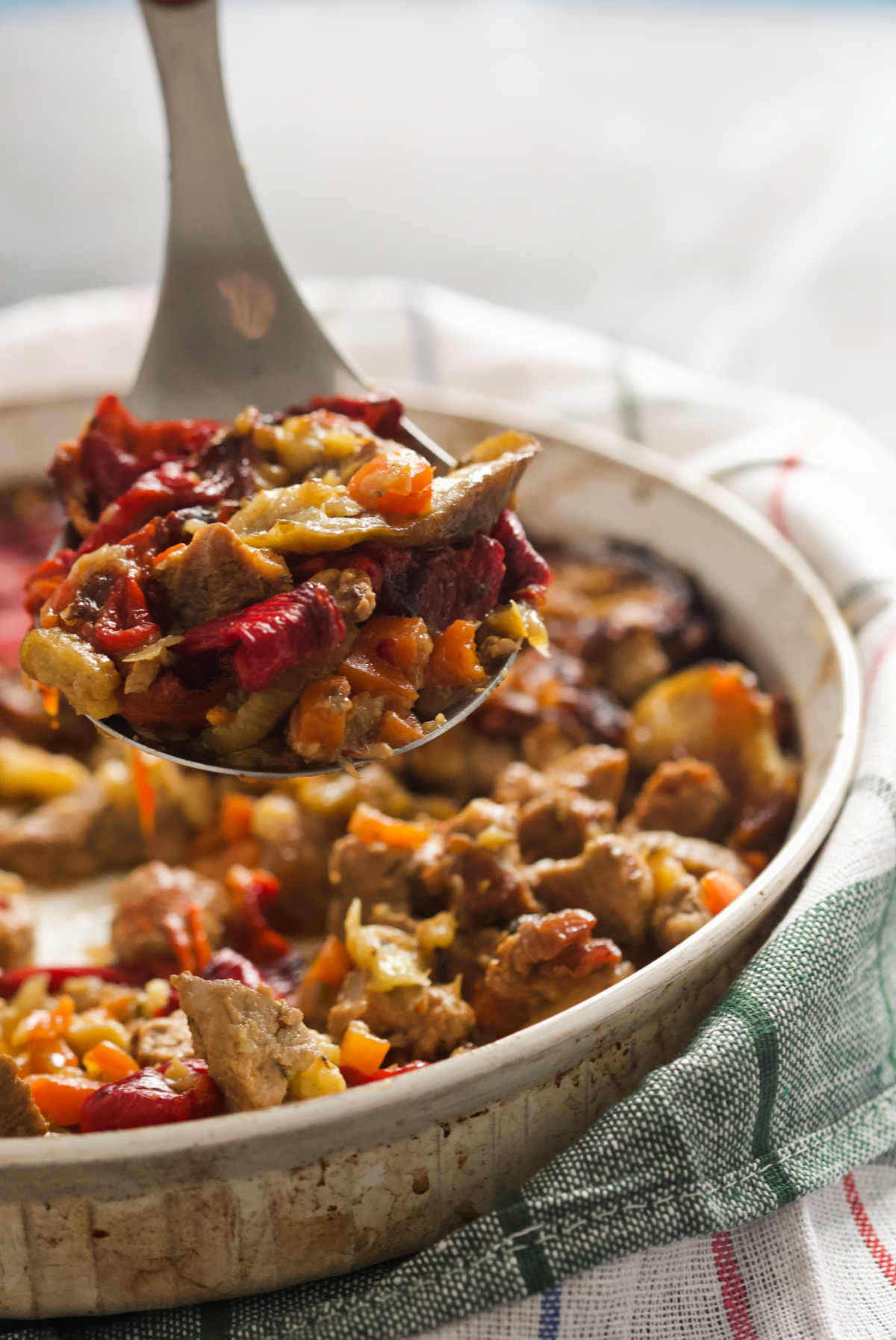 Round white casserole sitting on a kitchen towel, containing eggplant, red peppers, garlic and beef scooped on a silver spoon on a light background.