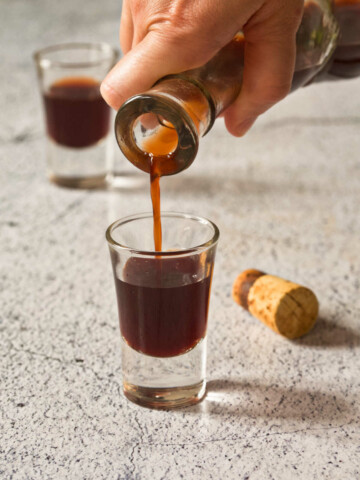 A hand holding a bottle and pouring liqueur into a shot glass on a gray background.