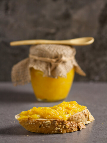 A piece of integral bread with marmalade and a jar of marmalade with a wooden spoon on top, on a gray background.